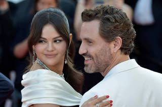 Selena Gomez and Edgar Ramirez pose on the red carpet, smiling; Selena is wearing an elegant gown with a shawl, and Edgar is in a suit