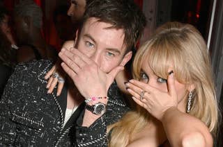 Barry Keoghan and Sabrina Carpenter pose on a red carpet event, covering their mouths with their hands. Barry wears a patterned shirt and black pants; Sabrina wears a fitted black dress