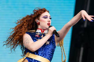 Chappell Roan with long curly hair on stage, wearing a sparkling blue bodysuit with gold accents, holding a microphone mid-performance