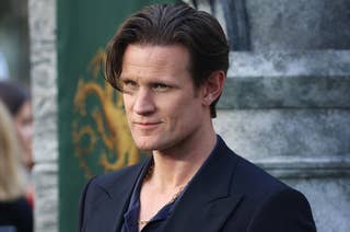 Matt Smith at an event, wearing a dark suit with a dark shirt, standing in front of a crowd and a stone structure