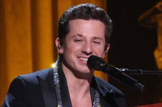 Charlie Puth sits at a red Yamaha piano on stage, wearing a formal suit with an embellished shirt while smiling and singing