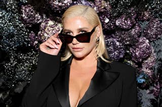 Kesha wearing sunglasses and a black jacket, posing in front of a floral backdrop