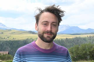 Kieran Culkin stands outdoors in front of a scenic landscape with hills and a pond, wearing a striped long-sleeve shirt