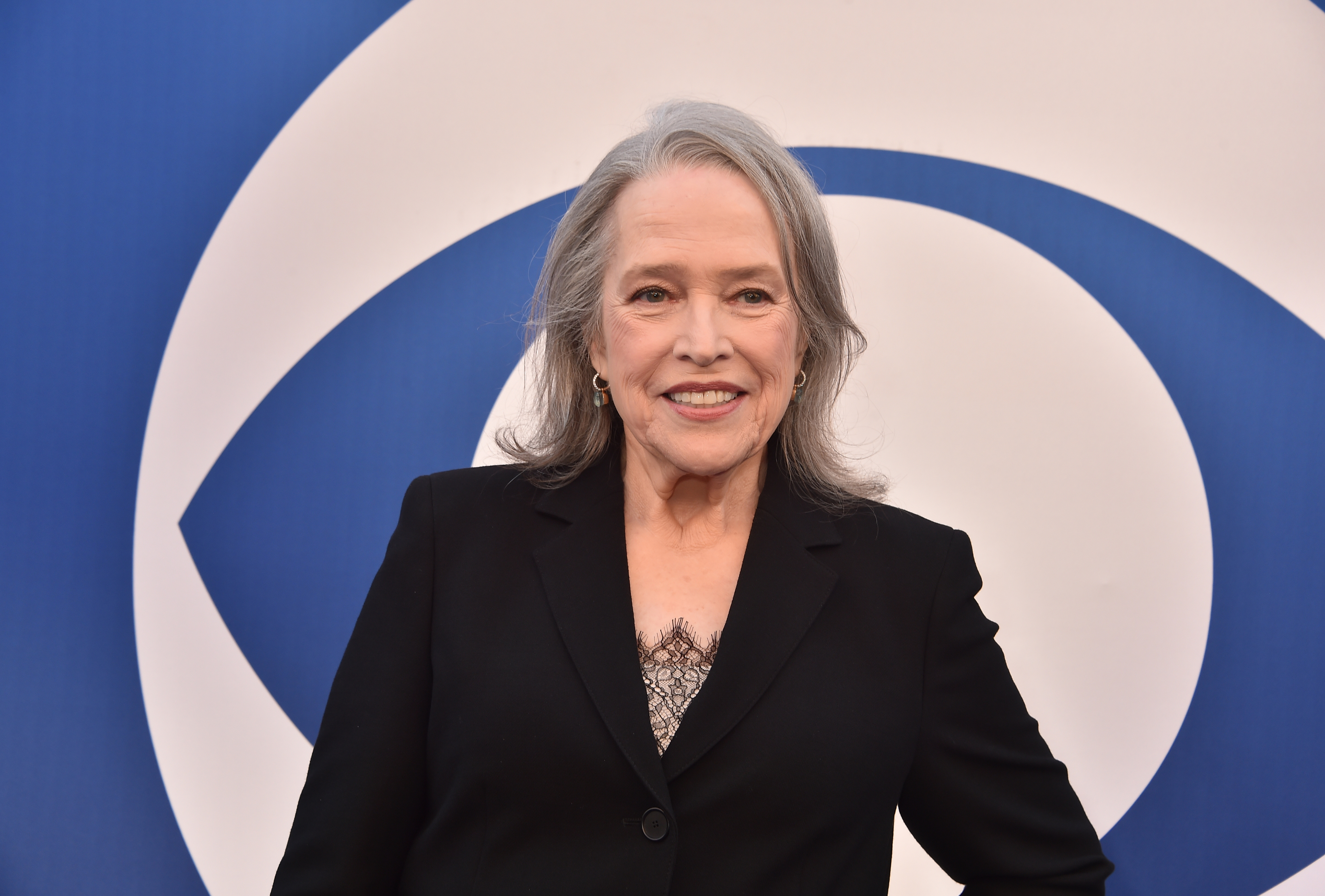 Kathy Bates with long gray hair smiles on a blue and white abstract background, wearing a black blazer