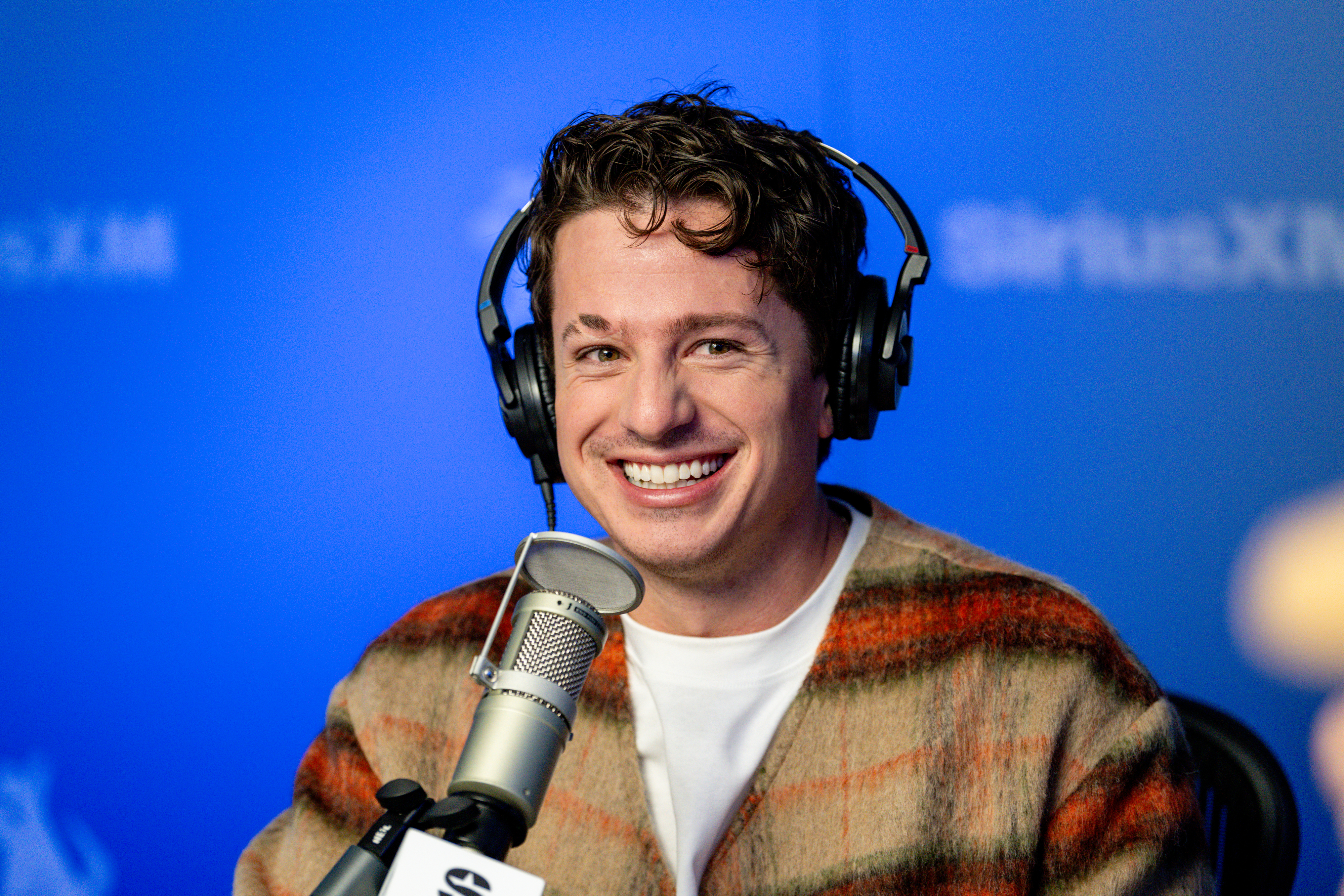 Charlie Puth smiling while wearing headphones and sitting in front of a microphone in a radio studio