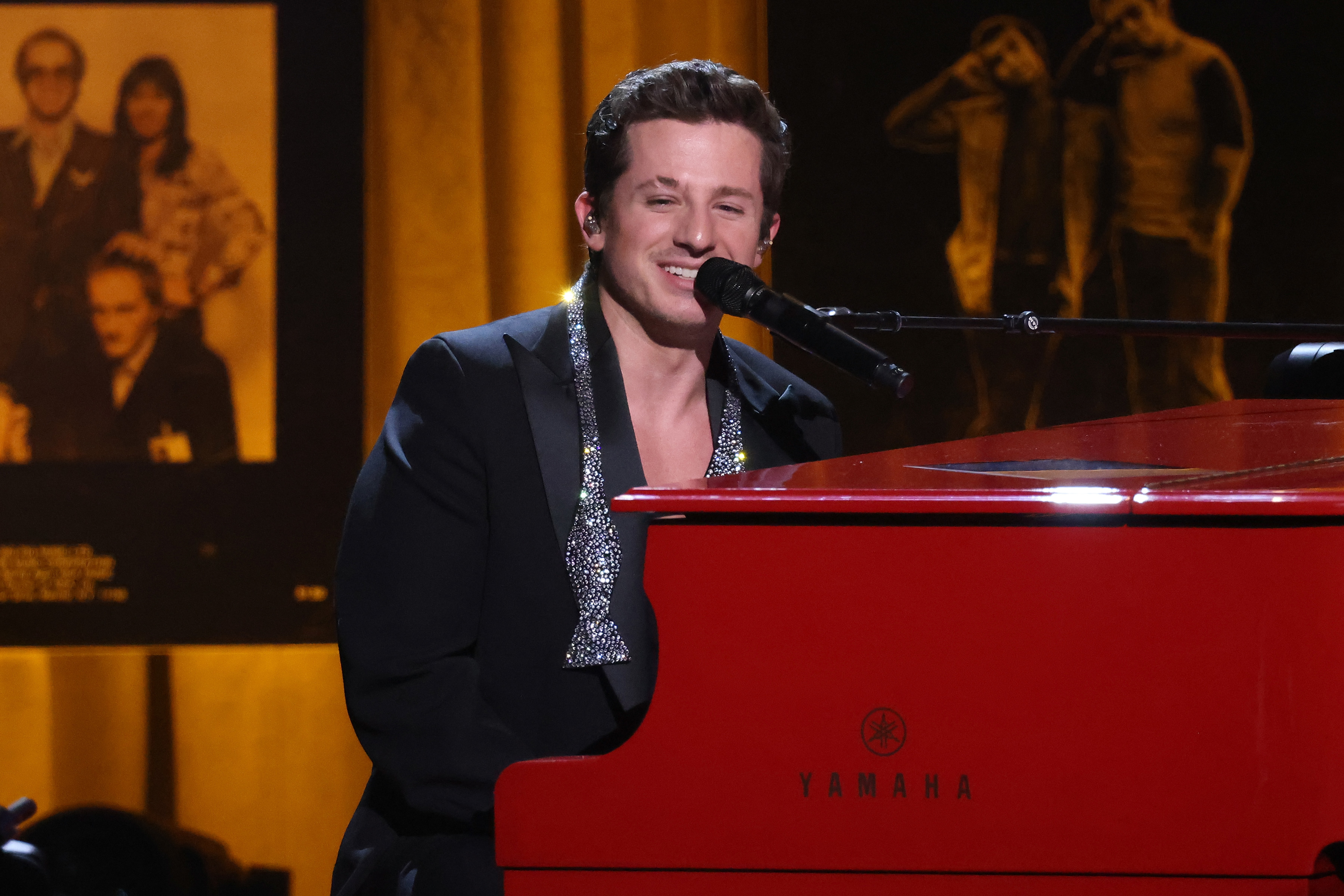 Charlie Puth sits at a Yamaha piano on stage, wearing a formal suit with an embellished shirt while smiling and singing