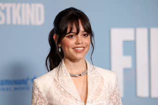 Jenna Ortega at a red carpet event, wearing a white lace blazer with a diamond necklace. She smiles for the cameras with a light blue background behind her