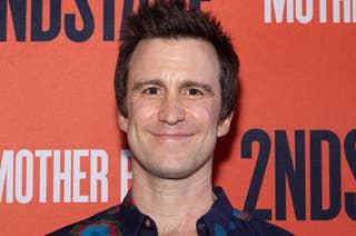 Gavin Creel stands in front of a red backdrop featuring the text 